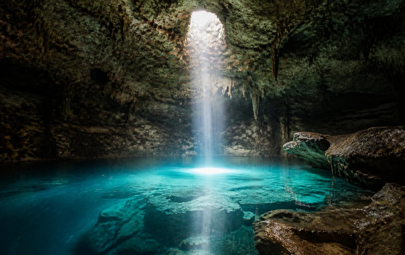 A single beam of daylight falling into a limestone cave onto still turquoise cenote water